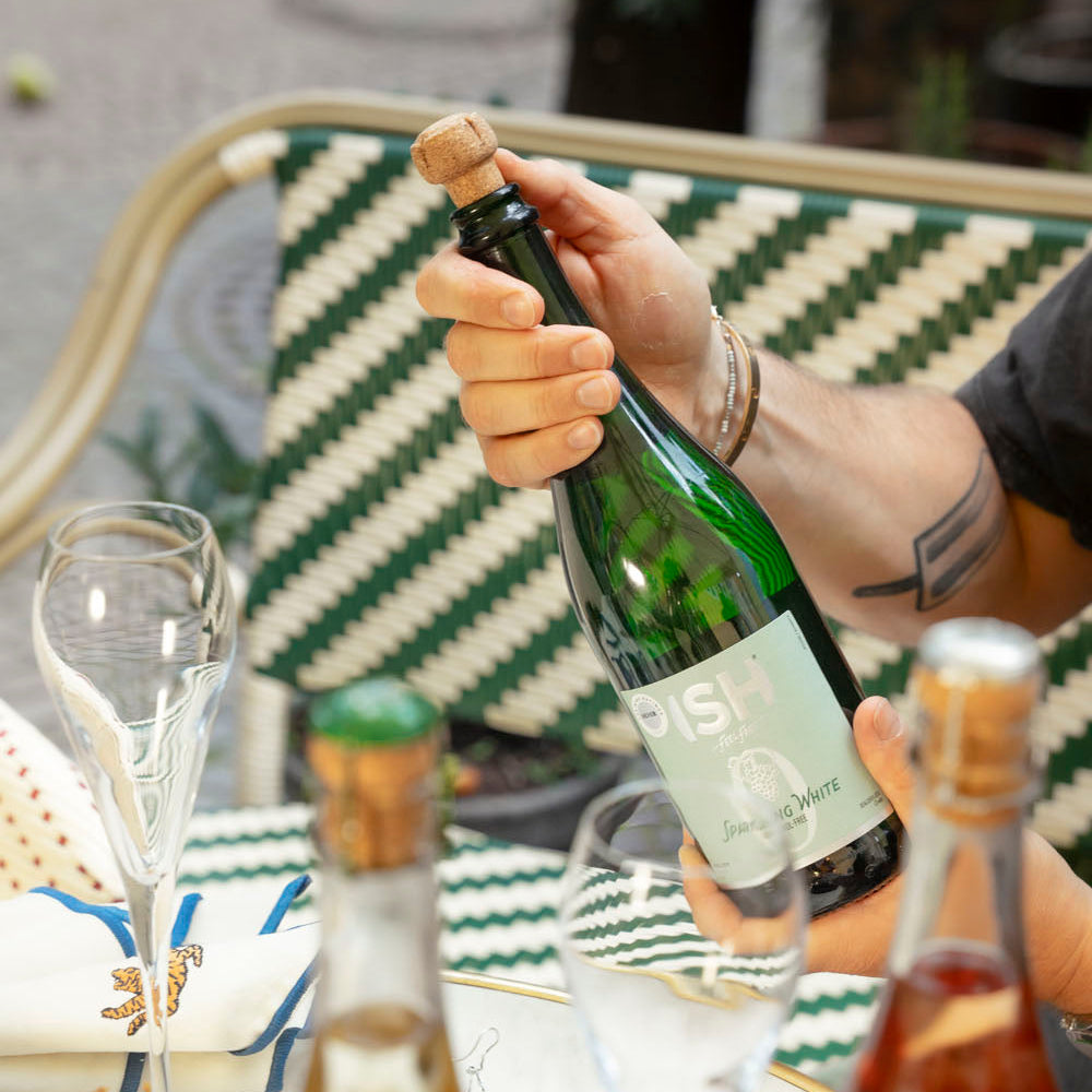 Person opening a bottle of alcohol-free champagne at a table with other bottles and glasses.
