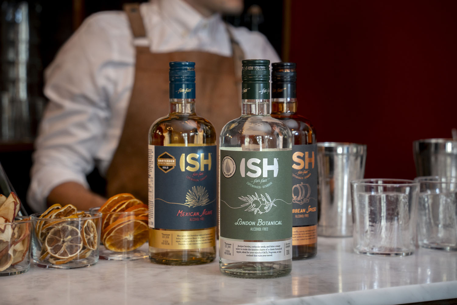 Bottles of 'ISH' brand alcohol-free spirits on a bar with a bartender in the background.